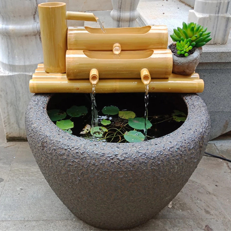 A decorative bamboo and stone water feature with a large round gray basin, a bamboo stream channel, small spouts with flowing water, and a small potted succulent, shown outdoors against a stone wall.