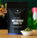 August Olympus Mint Cucumber Herbal Tea in a black resealable pouch, shown on a wooden surface with a small white bowl of loose herbal tea in front and a blurred indoor background.