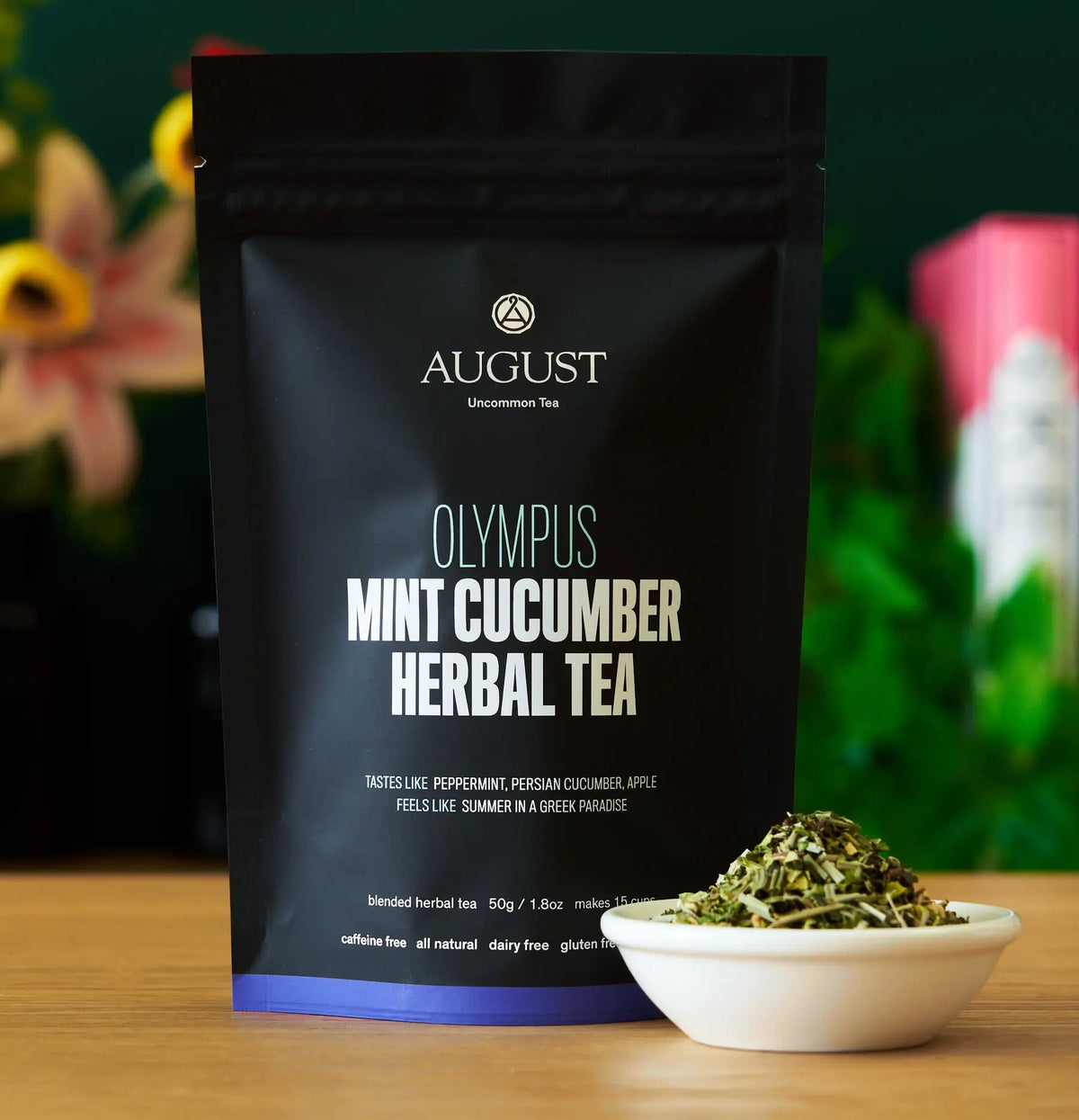 August Olympus Mint Cucumber Herbal Tea in a black resealable pouch, shown on a wooden surface with a small white bowl of loose herbal tea in front and a blurred indoor background.