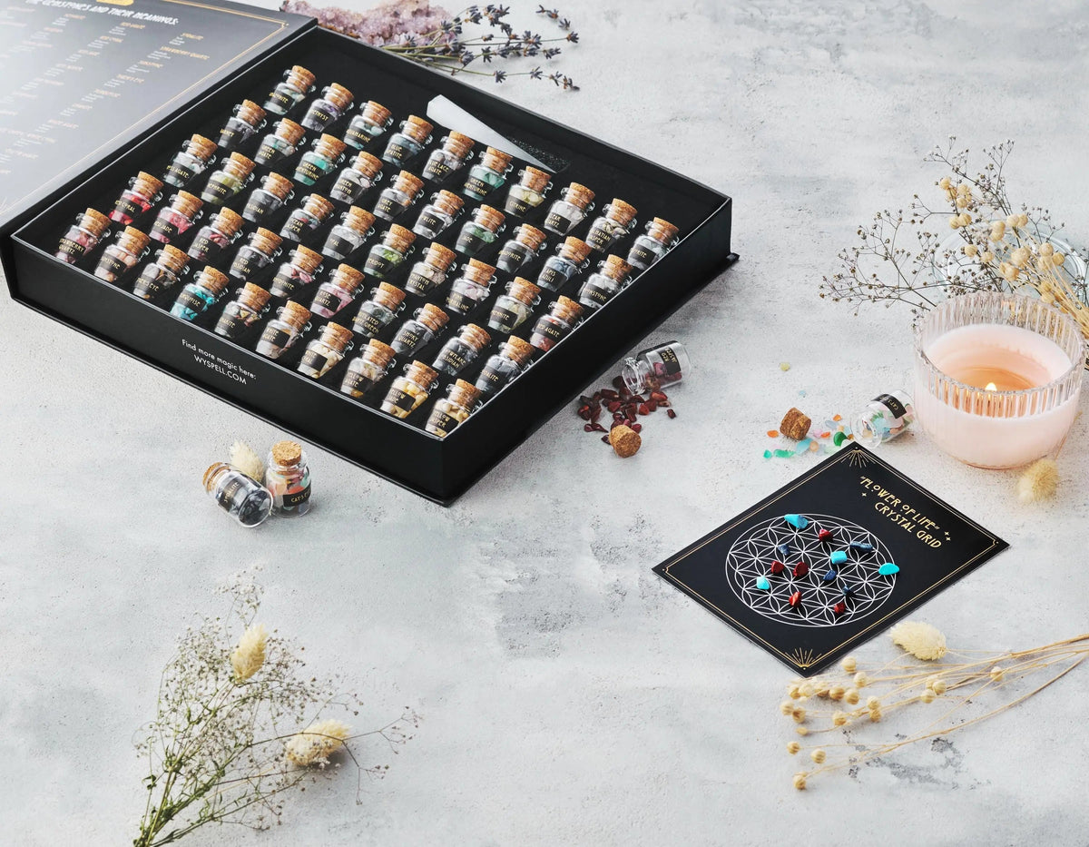 A 50 PCS Gemstone Kit featuring small corked glass bottles filled with various healing crystals, presented in an open black box next to a lit candle and a Flower of Life crystal grid card.