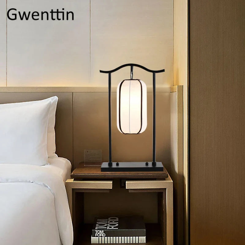 A modern table lamp with a black metal frame and a glowing white cylindrical shade, placed on a wooden bedside table next to a bed in a warm-toned bedroom. The brand name 'Gwenttin' is visible in the top left corner.
