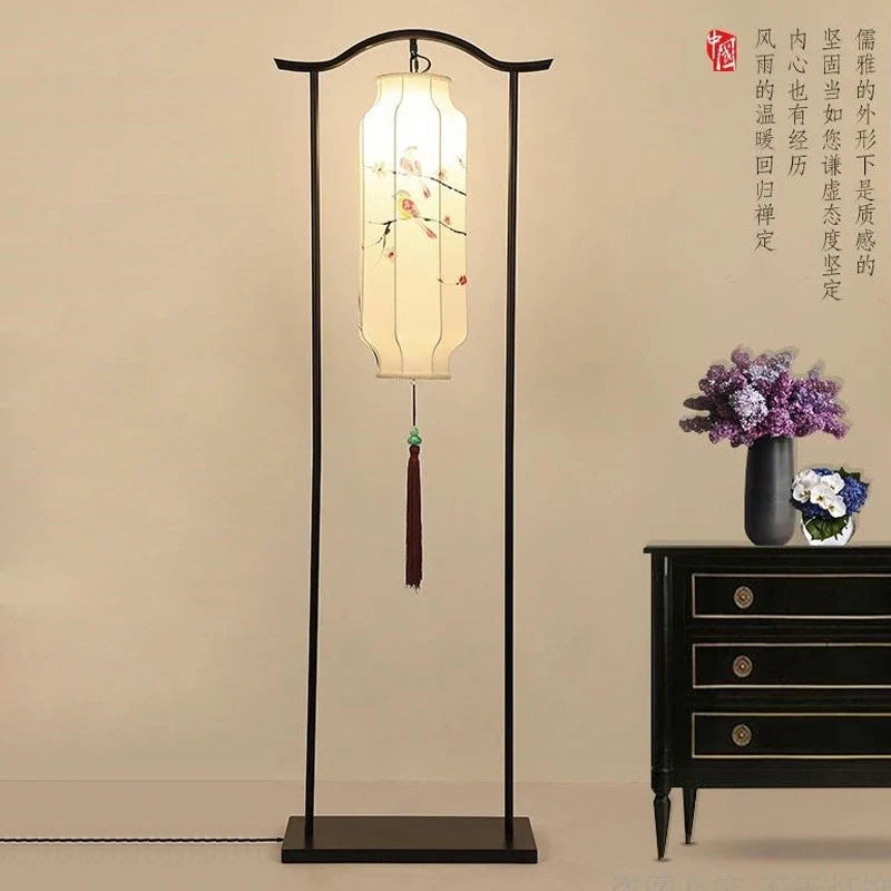 A traditional-style floor lamp with a black metal frame and a glowing rectangular white paper lantern featuring delicate floral illustrations. The lamp stands in a minimalist indoor setting next to a black wooden side table with purple flowers in a vase, against a warm beige wall with Chinese calligraphy.