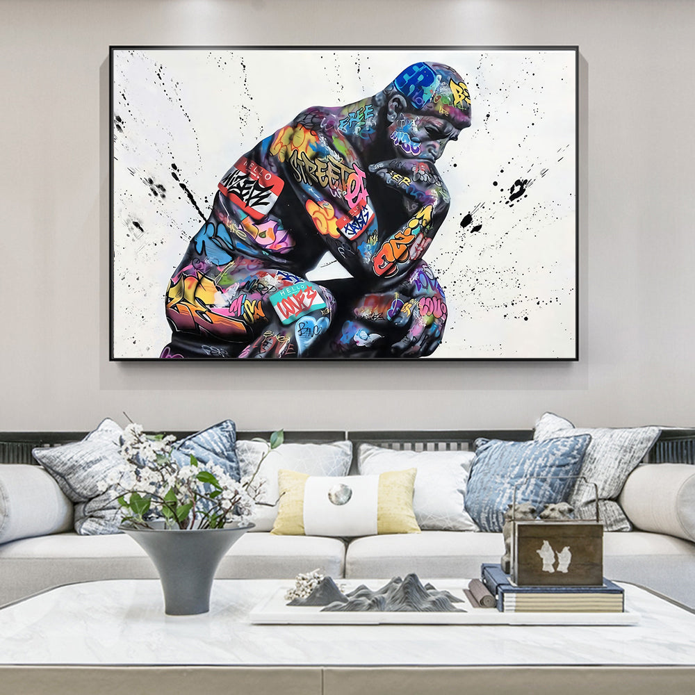 A large framed wall art print showing a colorful, paint-splattered seated figure on a white background, displayed above a modern sofa in a styled living room.