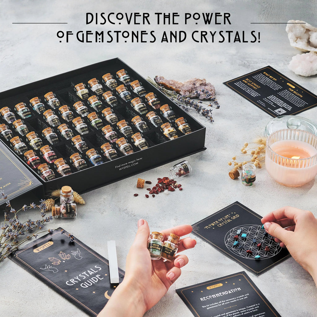 A 50 PCS Gemstone Kit featuring fifty labeled healing crystals in vials, with a pair of hands holding two, a crystal grid card, and information booklets arranged on a surface with a candle.