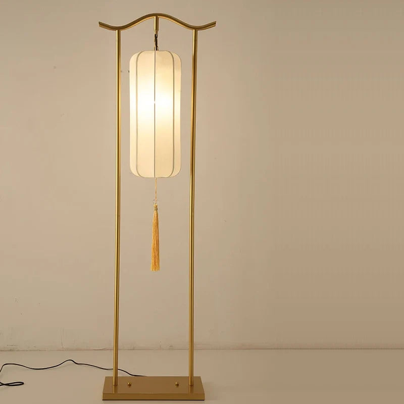 Gold floor lamp with a white shade and tassel against a beige wall.