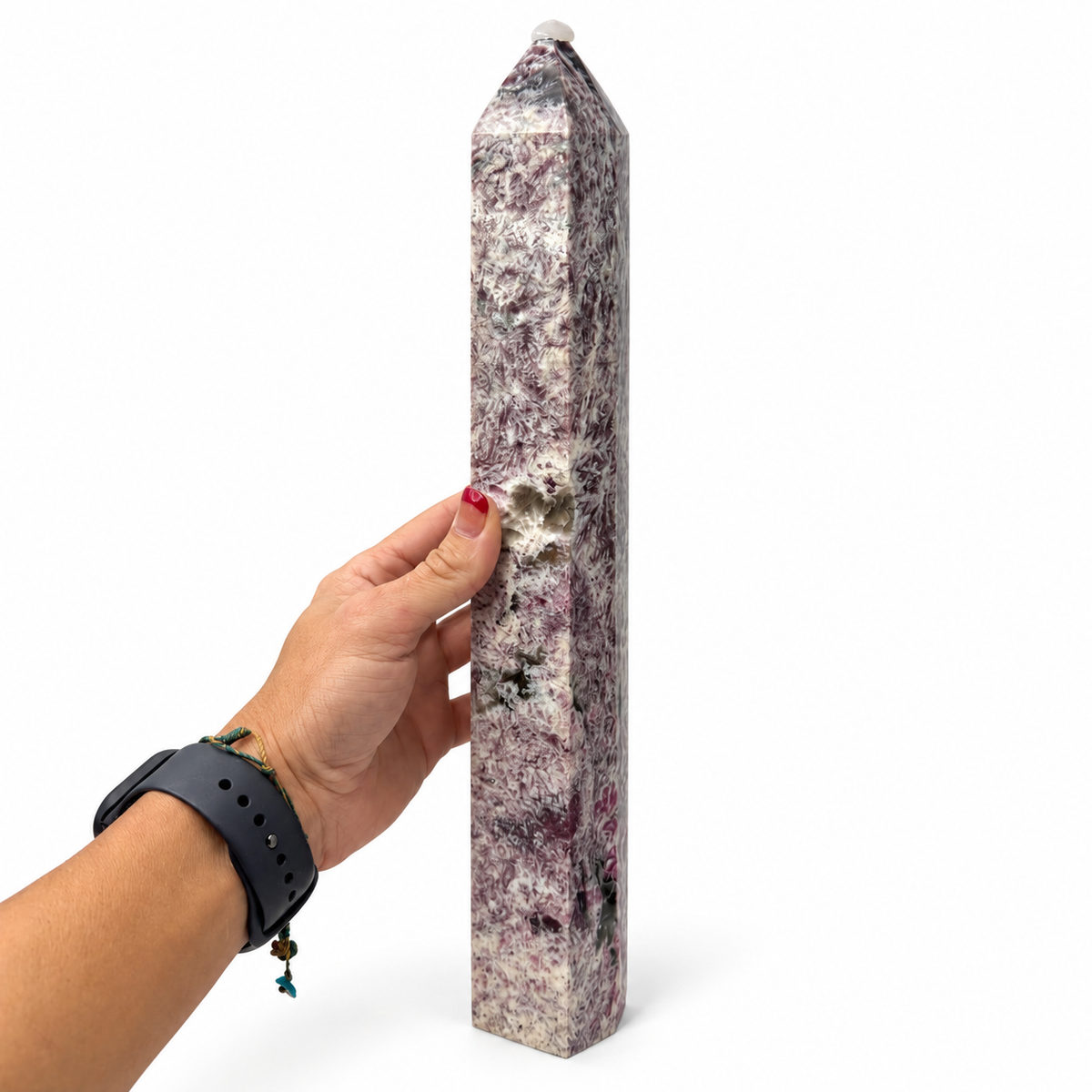A person's hand holds a tall, polished quartz crystal obelisk with natural pink, purple, and white marbled patterns against a plain white background.