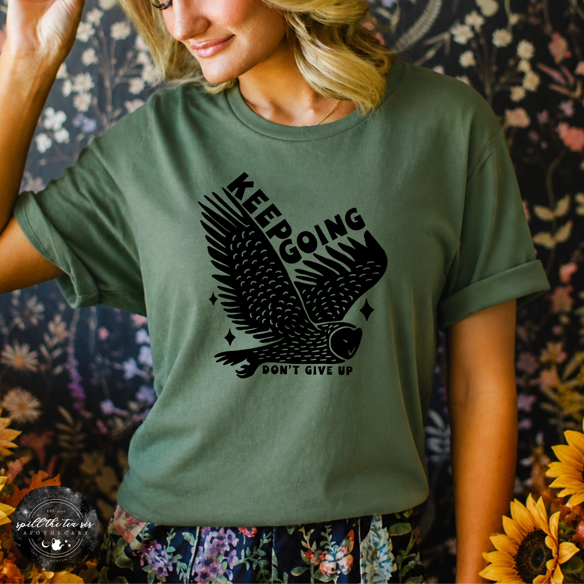A woman wearing an olive green graphic t-shirt with a black owl design and the text 'KEEPING DON'T GIVE UP'. The background is a dark floral pattern with sunflowers in the foreground.