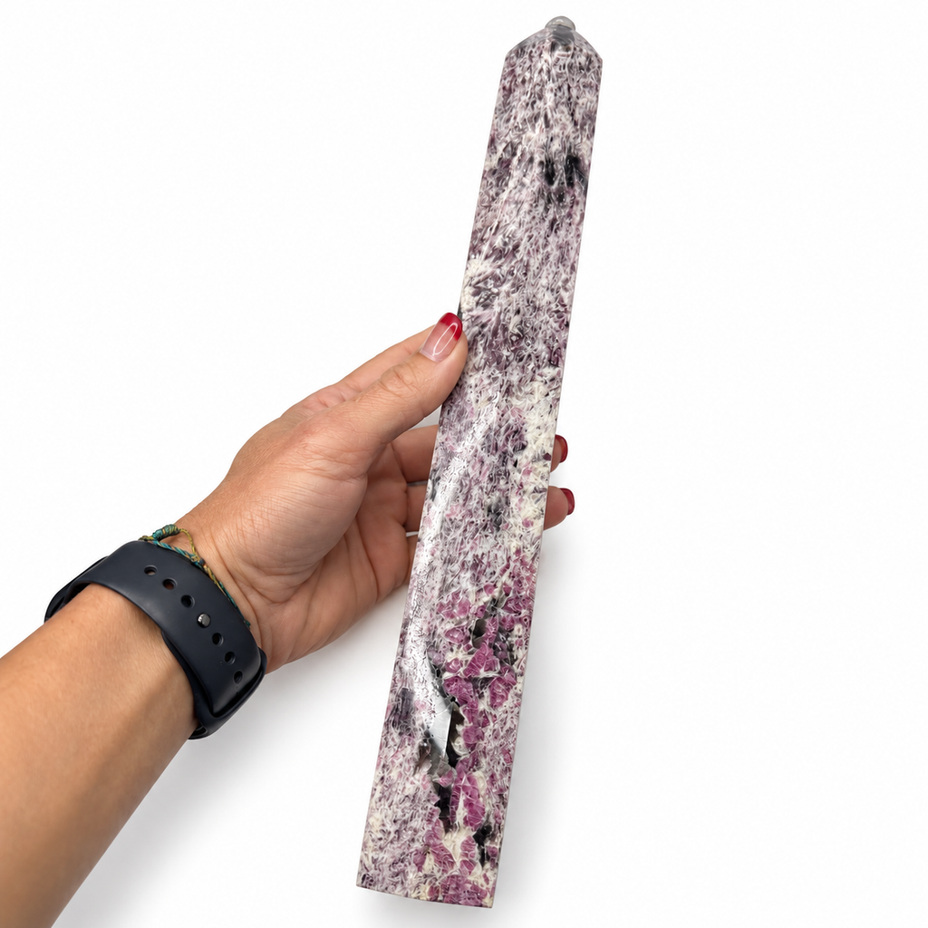 A hand holding a polished natural crystal tower, likely a quartz or calcite omen stone, showing a marbled pattern of purple, pink, gray, and white. The person holding it wears a black smartwatch on their wrist, set against a plain white background.