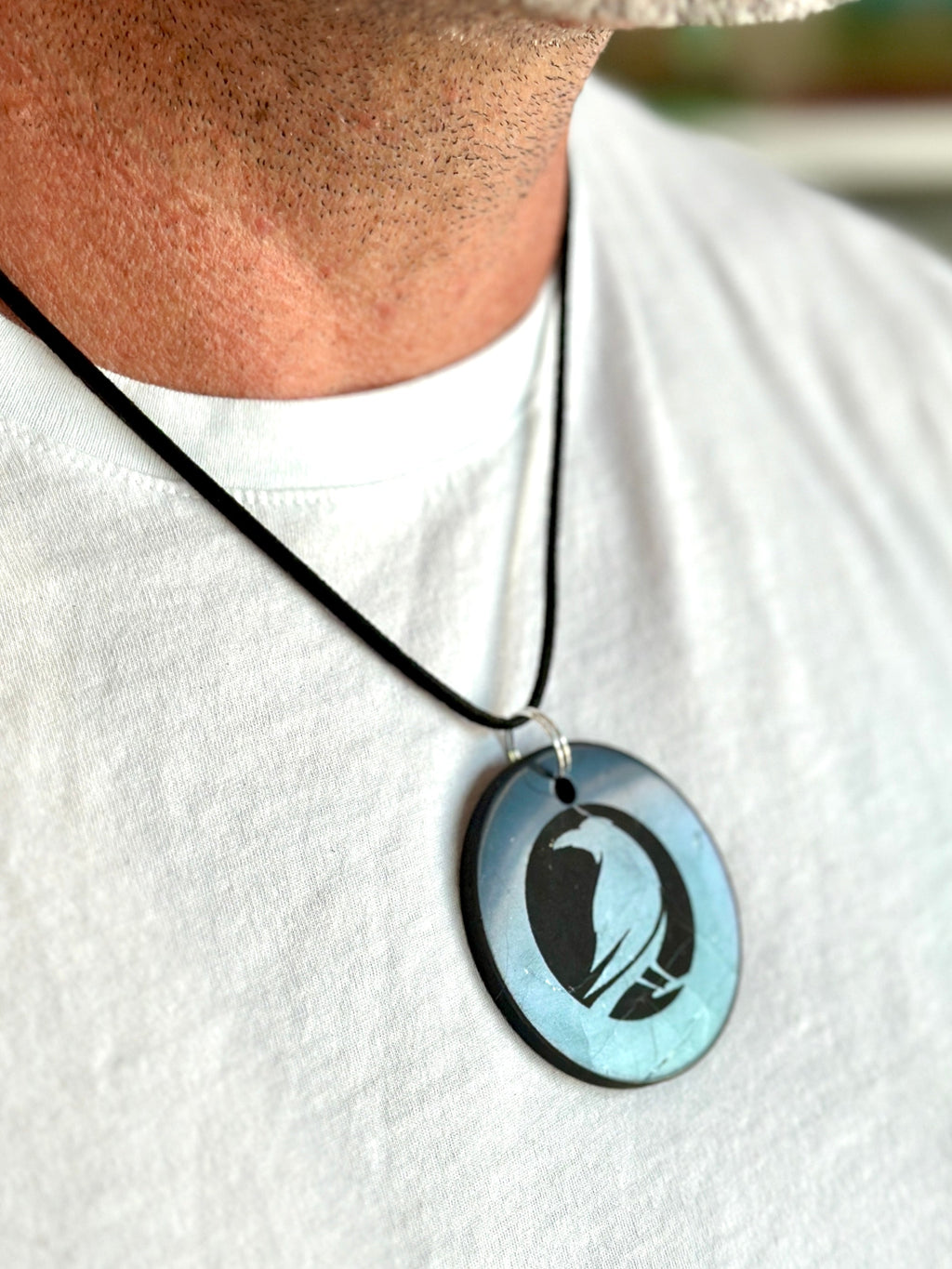 A close-up of a person wearing a black cord necklace with an oval pendant. The pendant has a light blue iridescent background with a black silhouette of a bird, likely a pelican or duck, and is shown against a white t-shirt.