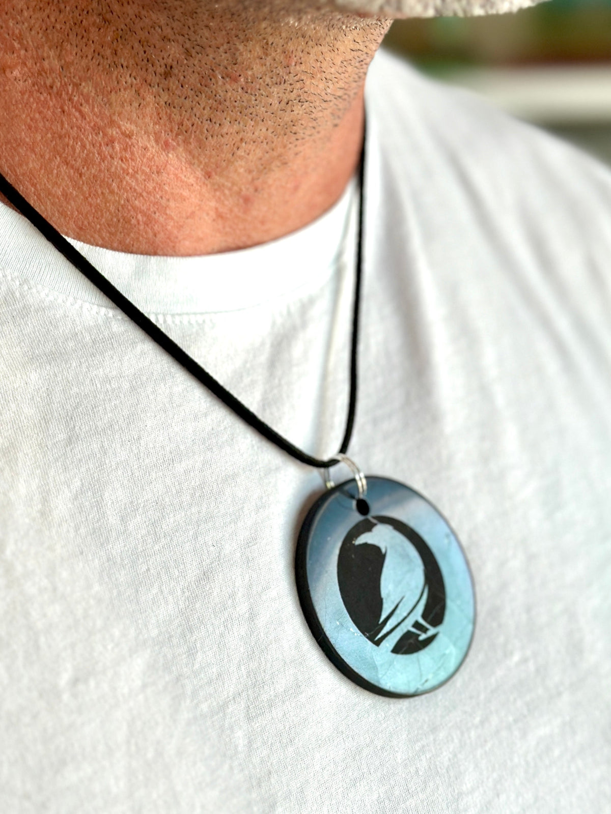 A close-up of a person wearing a black cord necklace with an oval pendant. The pendant has a light blue iridescent background with a black silhouette of a bird, likely a pelican or duck, and is shown against a white t-shirt.
