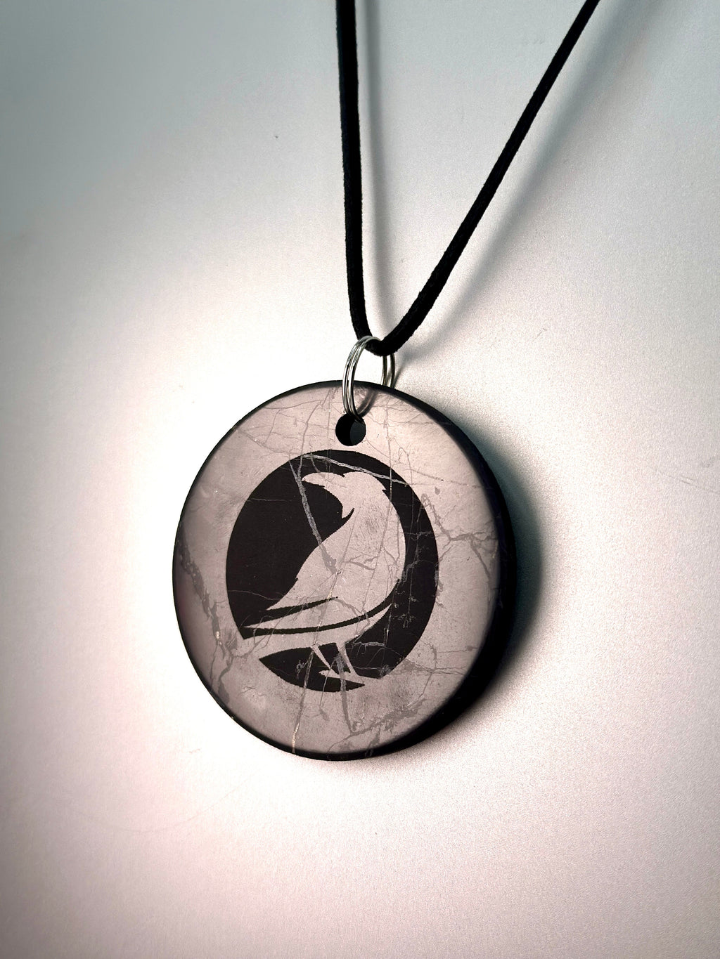 A round pendant necklace featuring a black bird silhouette over a cracked light gray background, suspended from a black cord with a silver ring.