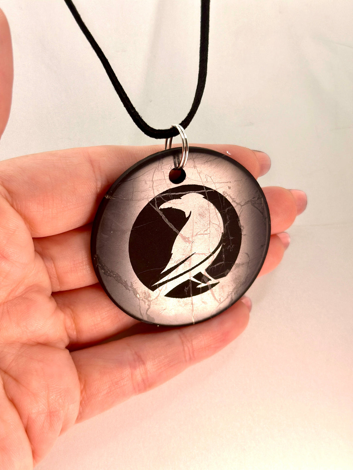 A hand holding a round pendant necklace with a black cord. The pendant features a black and white raven design on a cracked stone-like background.