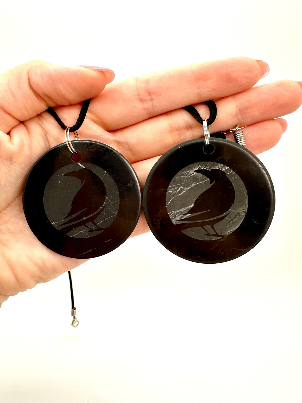 A hand holding two large round black pendant necklaces with black cords and silver jump rings, shown against a plain white background.