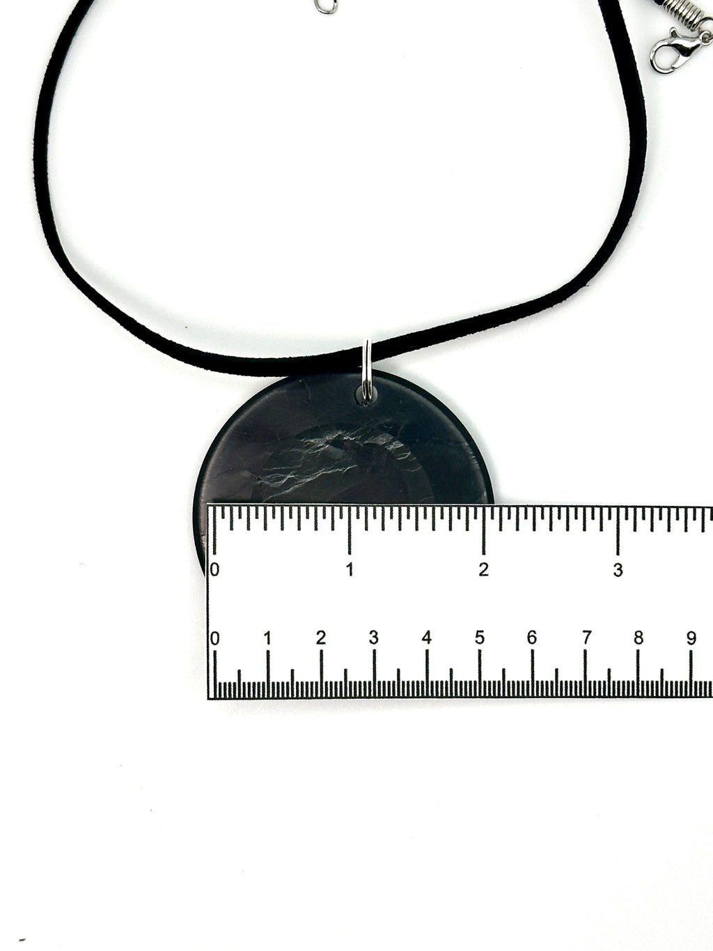 A black cord necklace with a round black pendant featuring a dark gray mountain design, shown with a metal ruler for scale on a white background.
