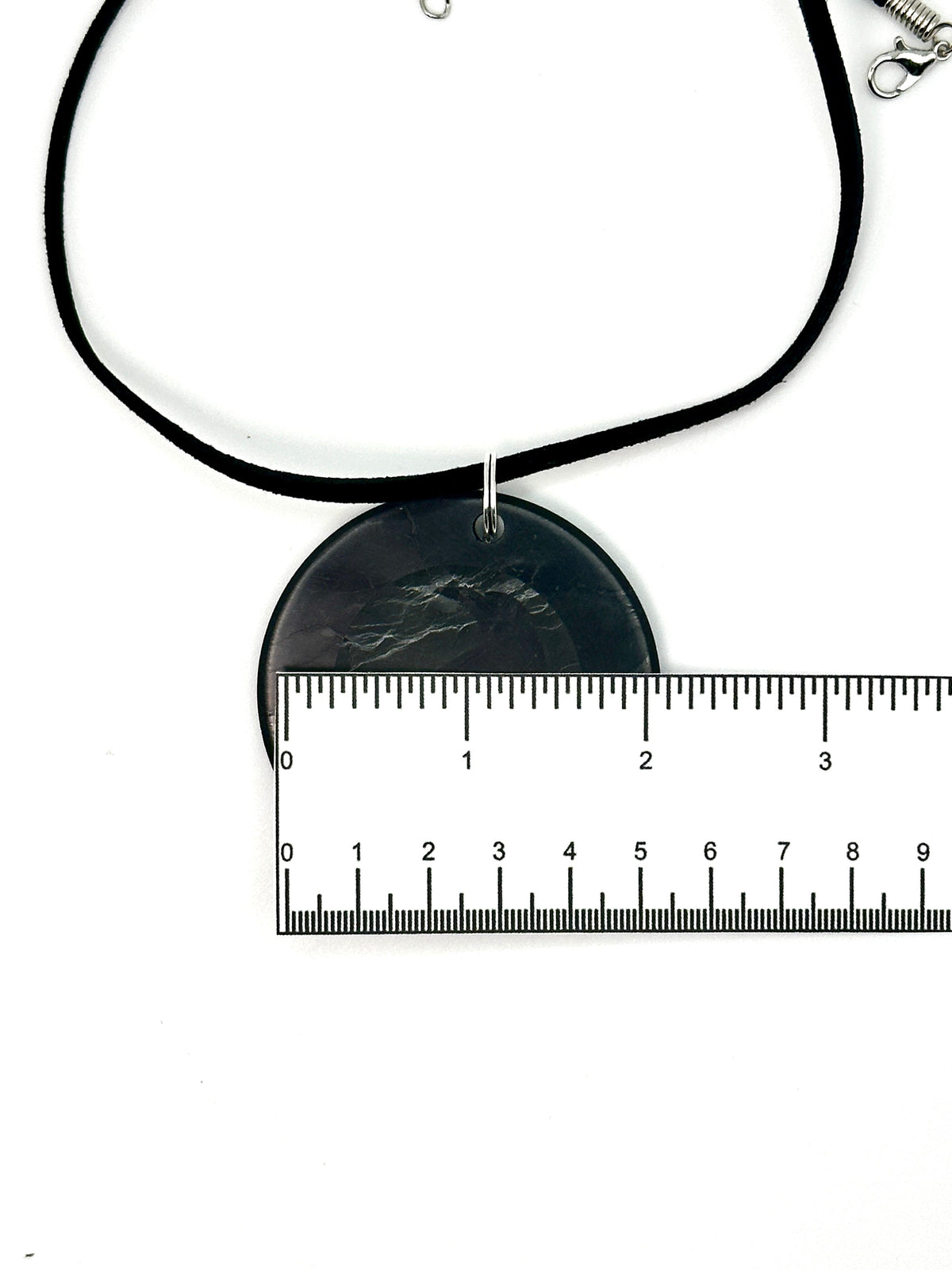 A black cord necklace with a round black pendant featuring a dark gray mountain design, shown with a metal ruler for scale on a white background.