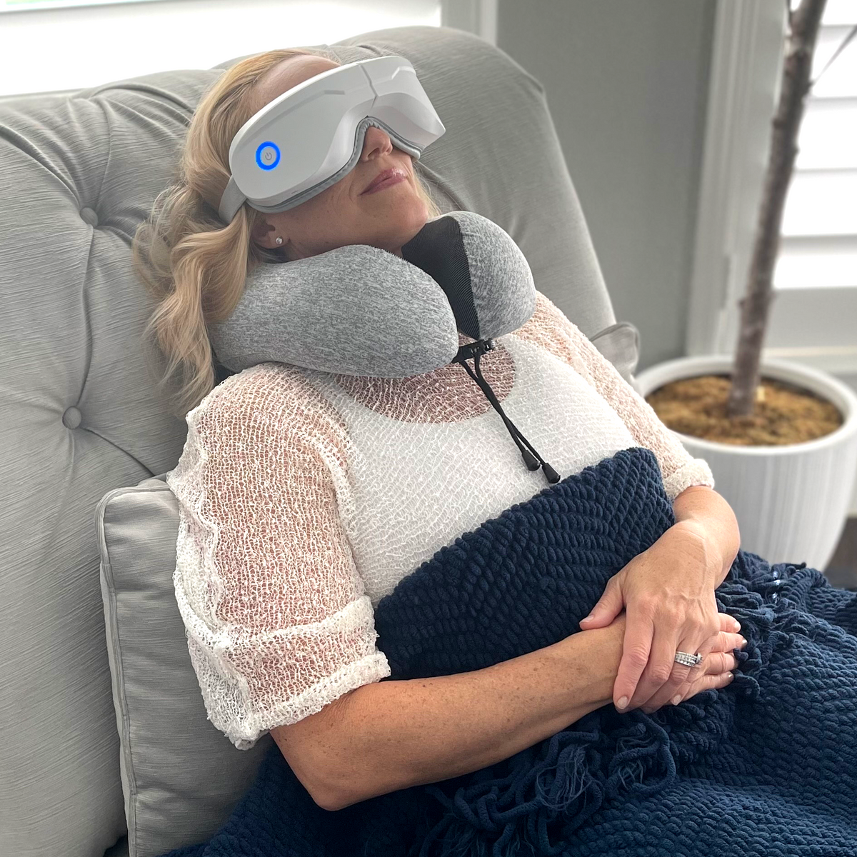 Woman relaxing on a couch with a neck pillow and eye mask, wrapped in a blanket.