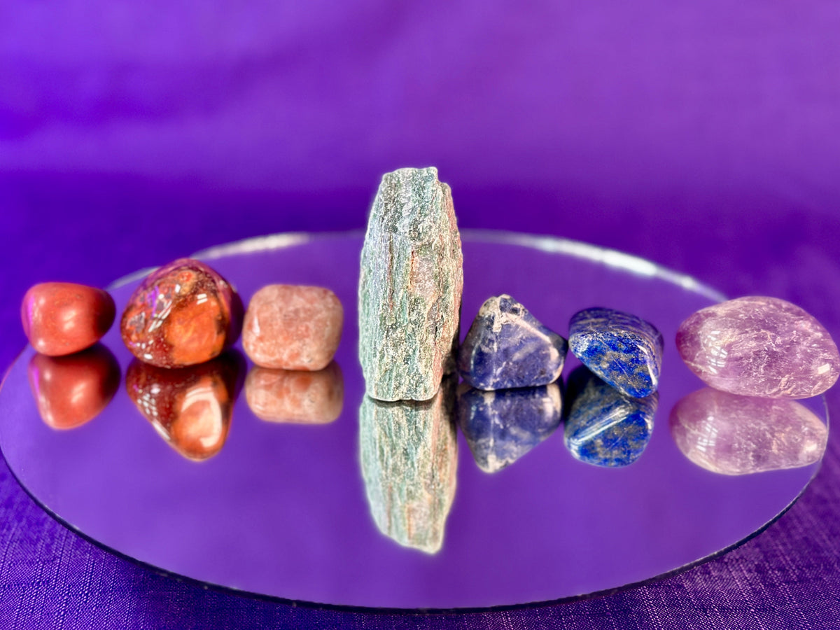 A set of polished and raw tumbled crystals arranged on a reflective circular mirror against a purple fabric background.