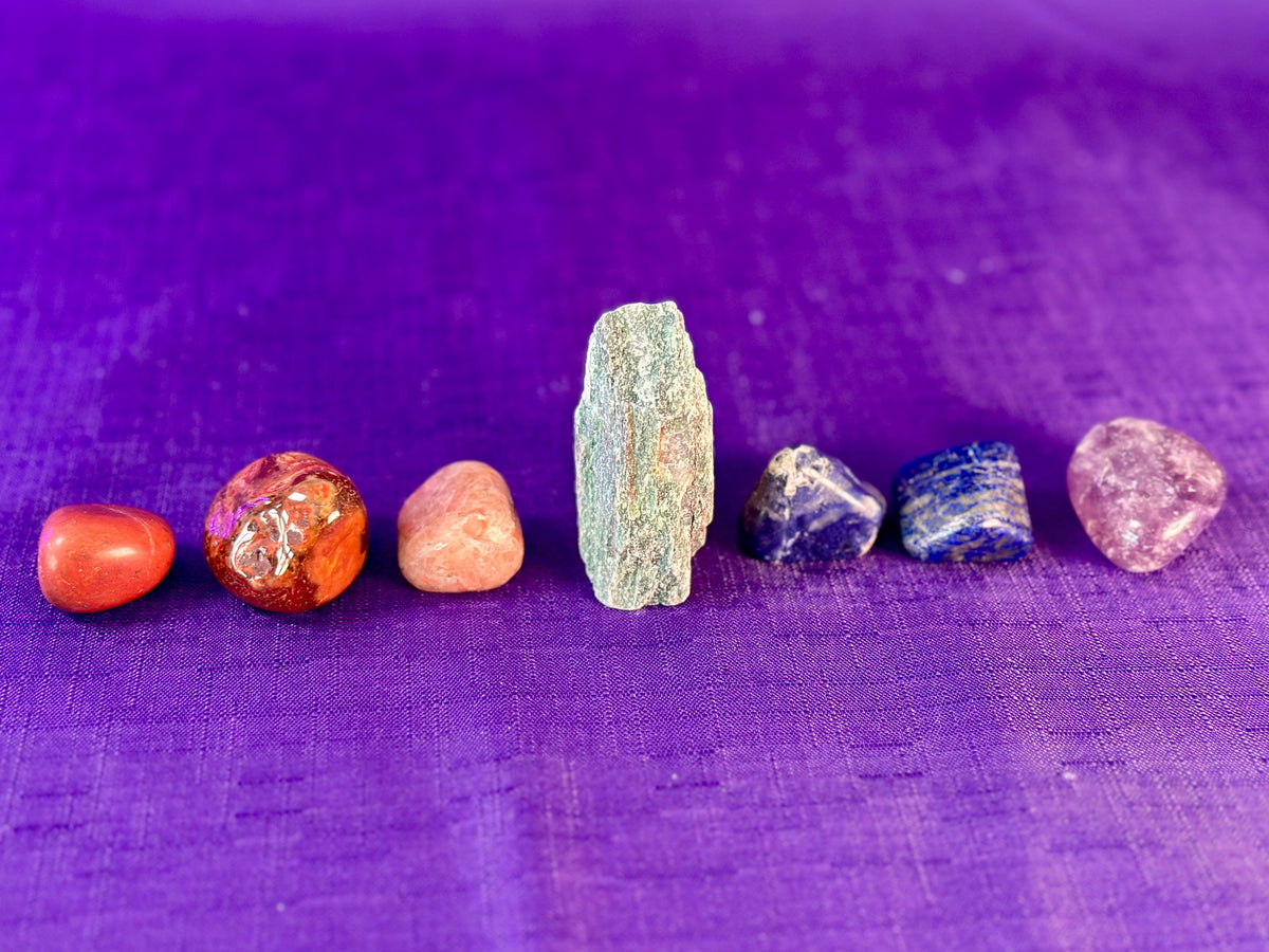 Seven polished and raw gemstones of various colors and textures arranged in a row on a textured purple fabric background.