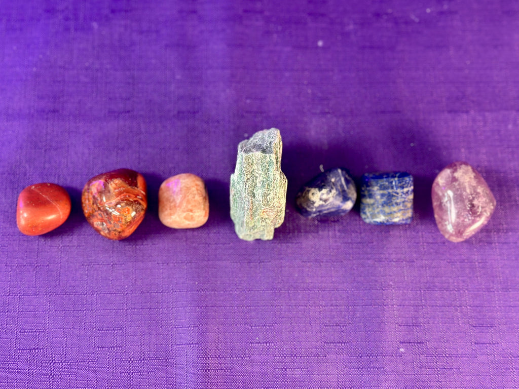 Seven raw, polished tumbled gemstones and a rough crystal cluster arranged in a row on a textured purple fabric background.