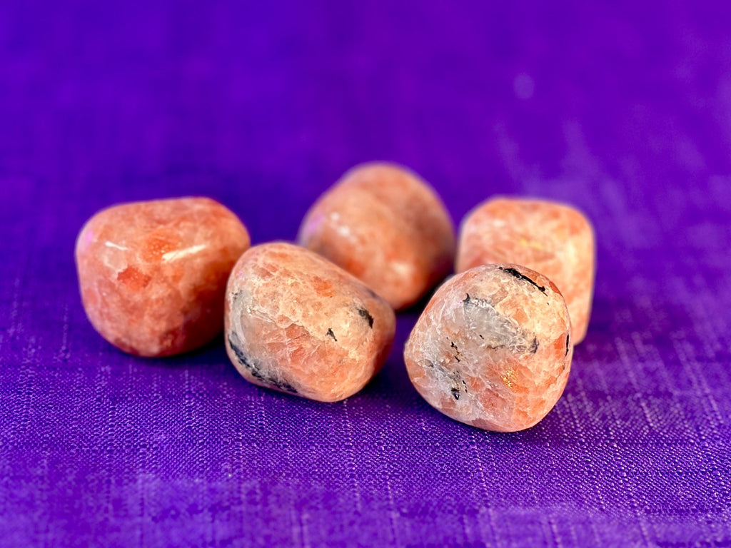 Five polished tumbled orange stone pendants arranged on a textured purple fabric background.