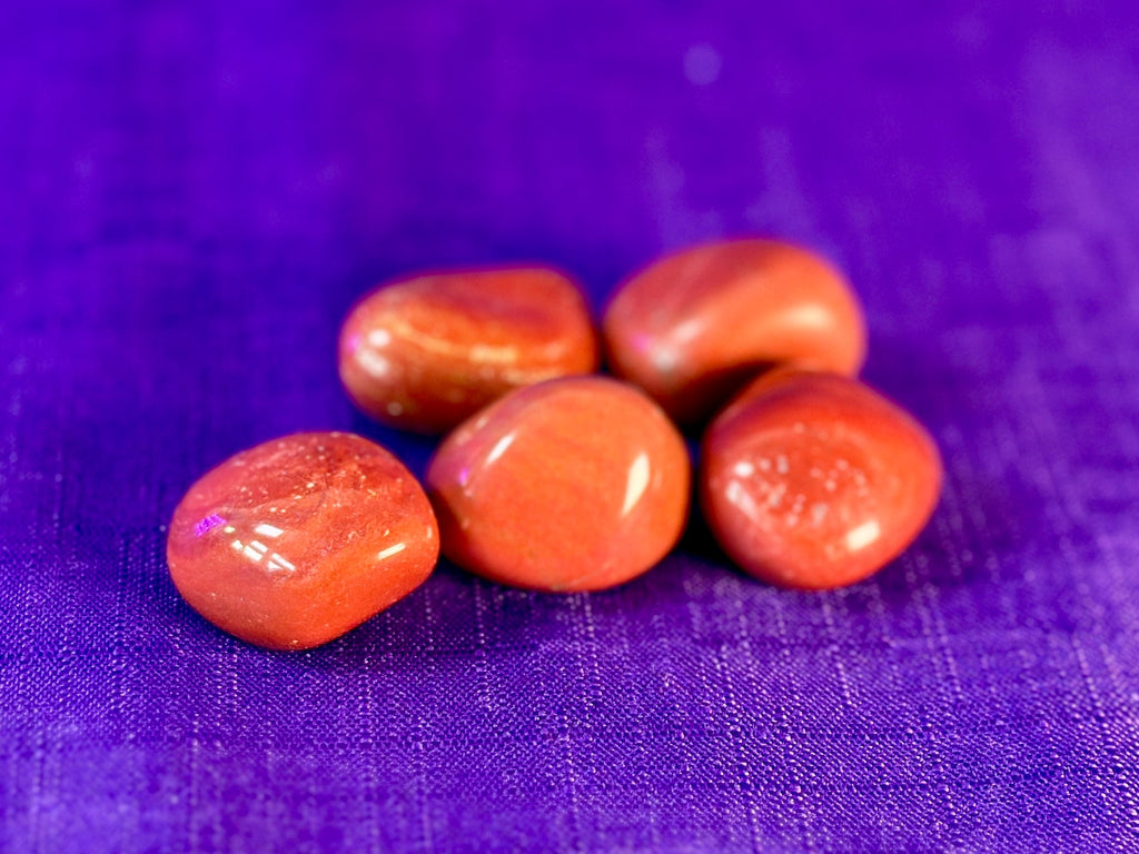 Five smooth, polished reddish-orange tumbled stones arranged on a textured purple fabric background.