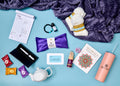 A curated gift box flat lay on a light blue background, featuring a purple plush blanket, white socks, a purple eye mask, a daily journal, a black pen in a black case, a small bottle, a white teapot, colorful tea packets, a pink tumbler, and other wellness-themed items.
