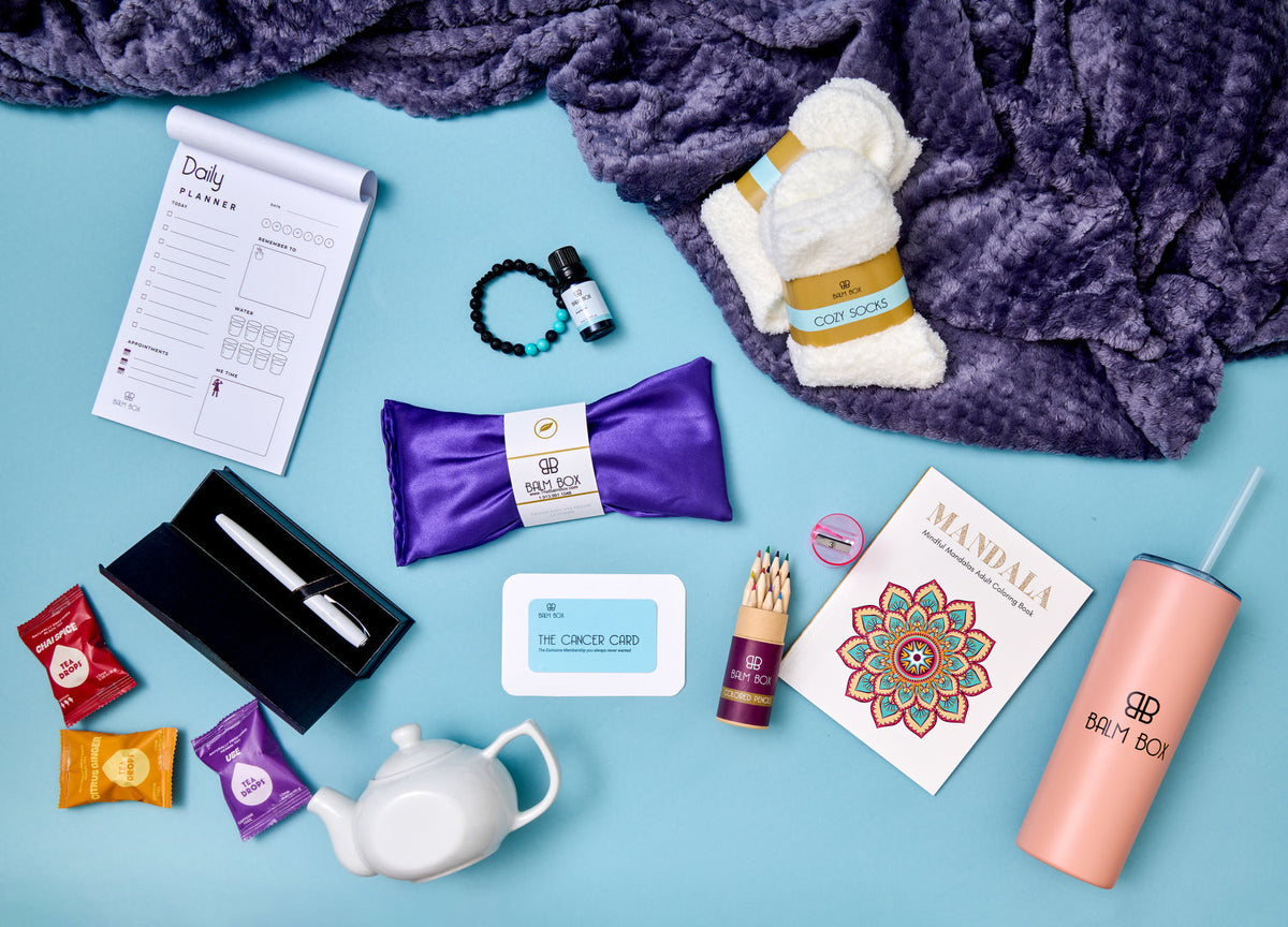 A curated gift box flat lay on a light blue background, featuring a purple plush blanket, white socks, a purple eye mask, a daily journal, a black pen in a black case, a small bottle, a white teapot, colorful tea packets, a pink tumbler, and other wellness-themed items.