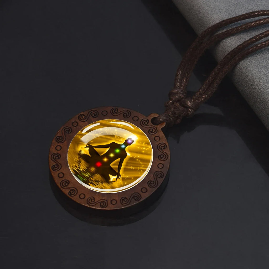 A round pendant necklace with a dark brown carved wooden frame and a brown cord, featuring a glowing yellow chakra design with red and green lights in the center, displayed on a dark reflective surface.