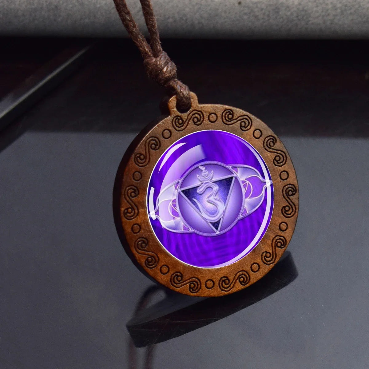A round pendant necklace with a brown engraved wooden frame and a purple center featuring a white Om symbol, hanging on a dark reflective surface.