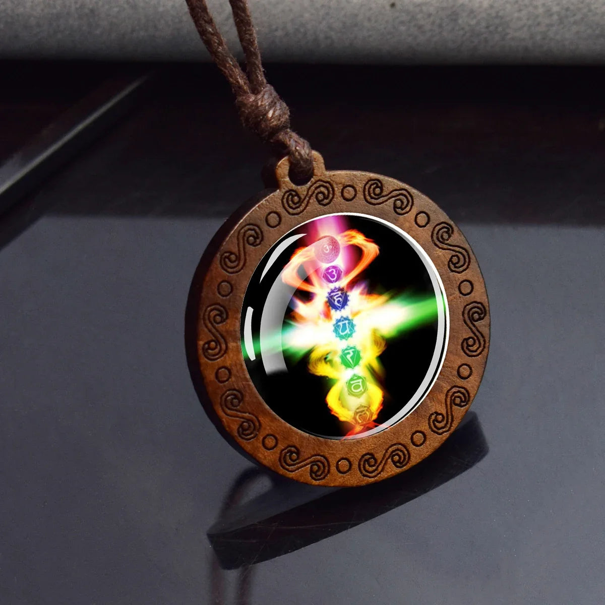 A round wooden pendant necklace with a carved brown spiral border, featuring a colorful chakra symbol on a black background, suspended from a brown cord.