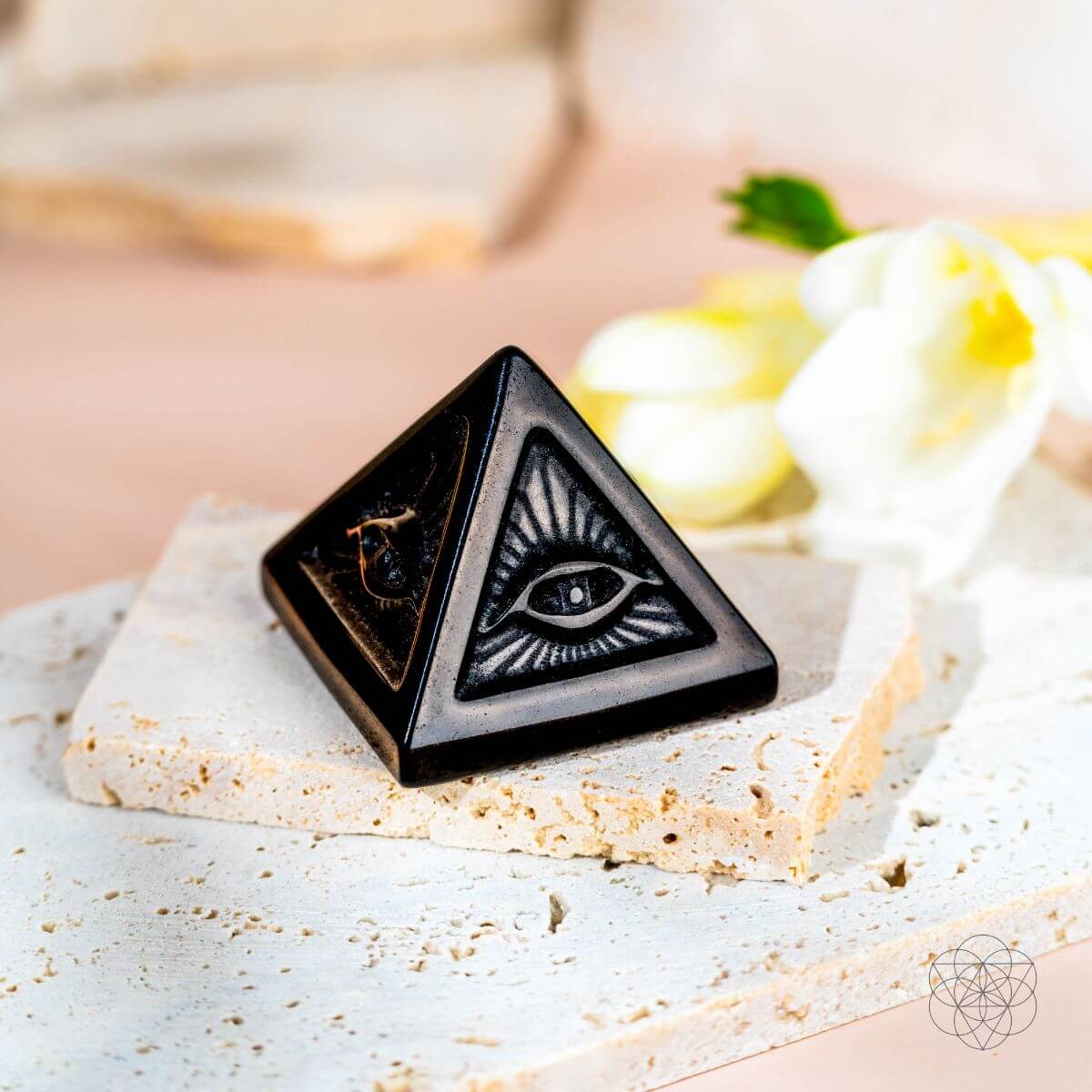 Black pyramid-shaped object with eye design on a textured surface with flowers in the background