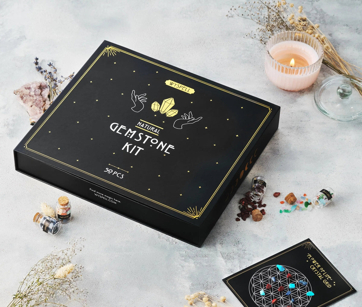 A black box labeled 50 PCS Gemstone Kit surrounded by small corked glass vials of healing crystals, a lit candle, dried flowers, and a printed crystal grid card.