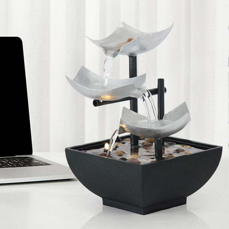 Decorative indoor water fountain with three tiered bowls on a white surface next to a laptop.