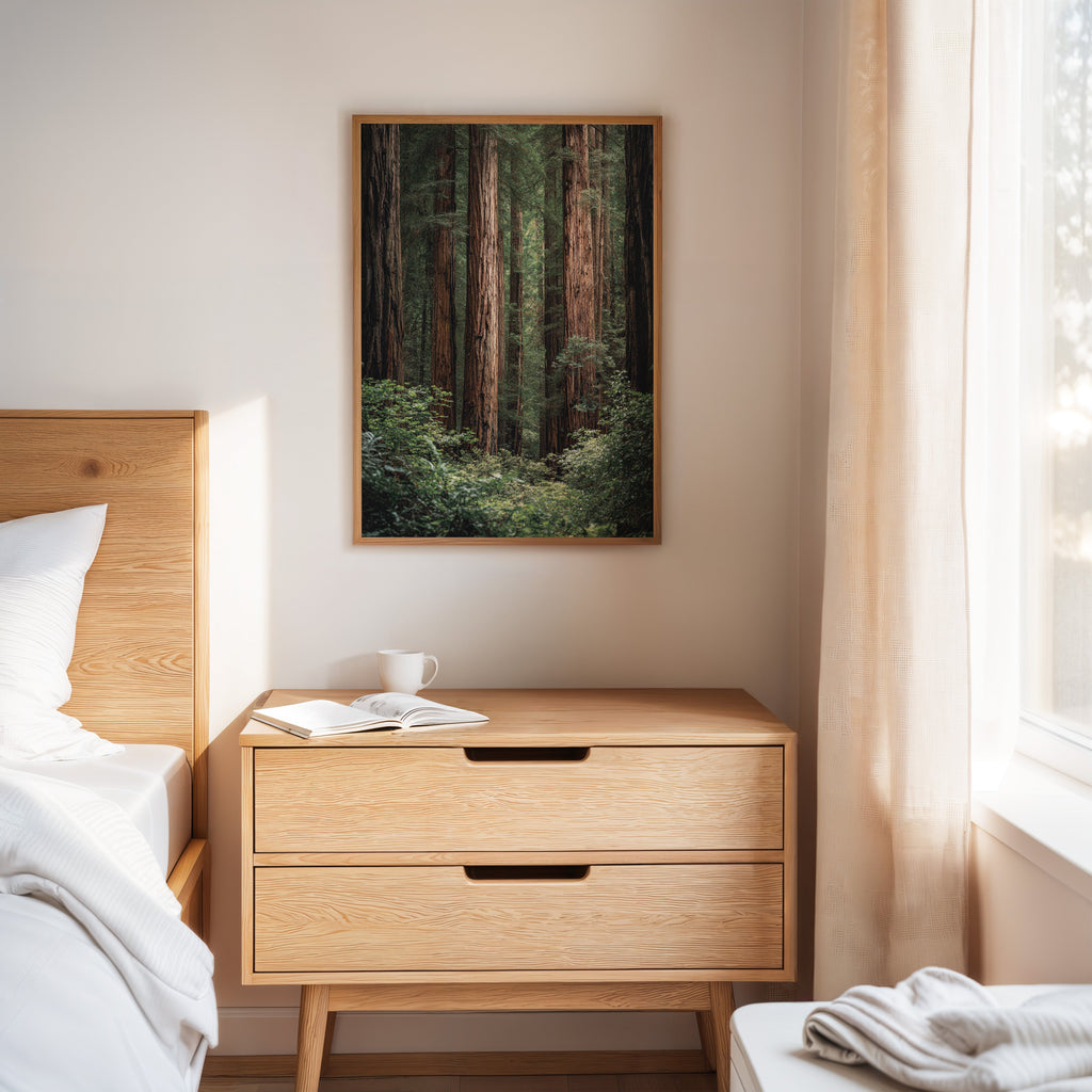 Wooden nightstand with a book and mug next to a bed, with a framed artwork above on a white wall.