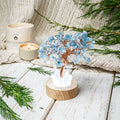 A decorative crystal tree centerpiece with light blue gemstone leaves and copper wire branches, displayed on a round wooden base with white crystal points. The setup is styled on a white wooden surface with evergreen branches, a cream knit blanket, and a lit candle in the background.