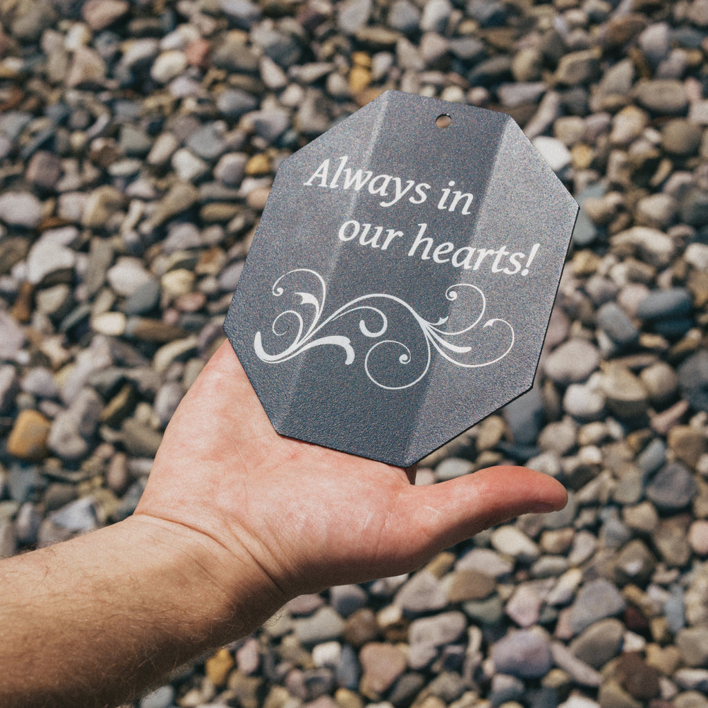 Hand holding a gray tag with 'Always in our hearts!' text against a pebbled background
