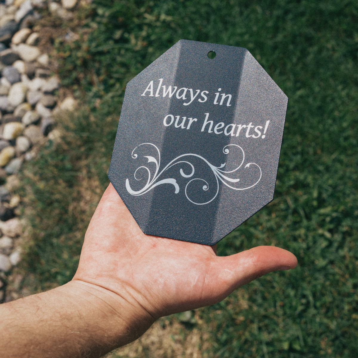 Hand holding a gray tag with 'Always in our hearts!' text against a natural background.