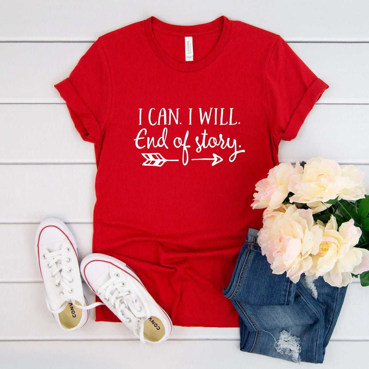 A red short-sleeve graphic T-shirt laid flat on a white wood background, styled with white sneakers, blue denim shorts, and pale pink flowers. The shirt has white text reading 'I CAN.