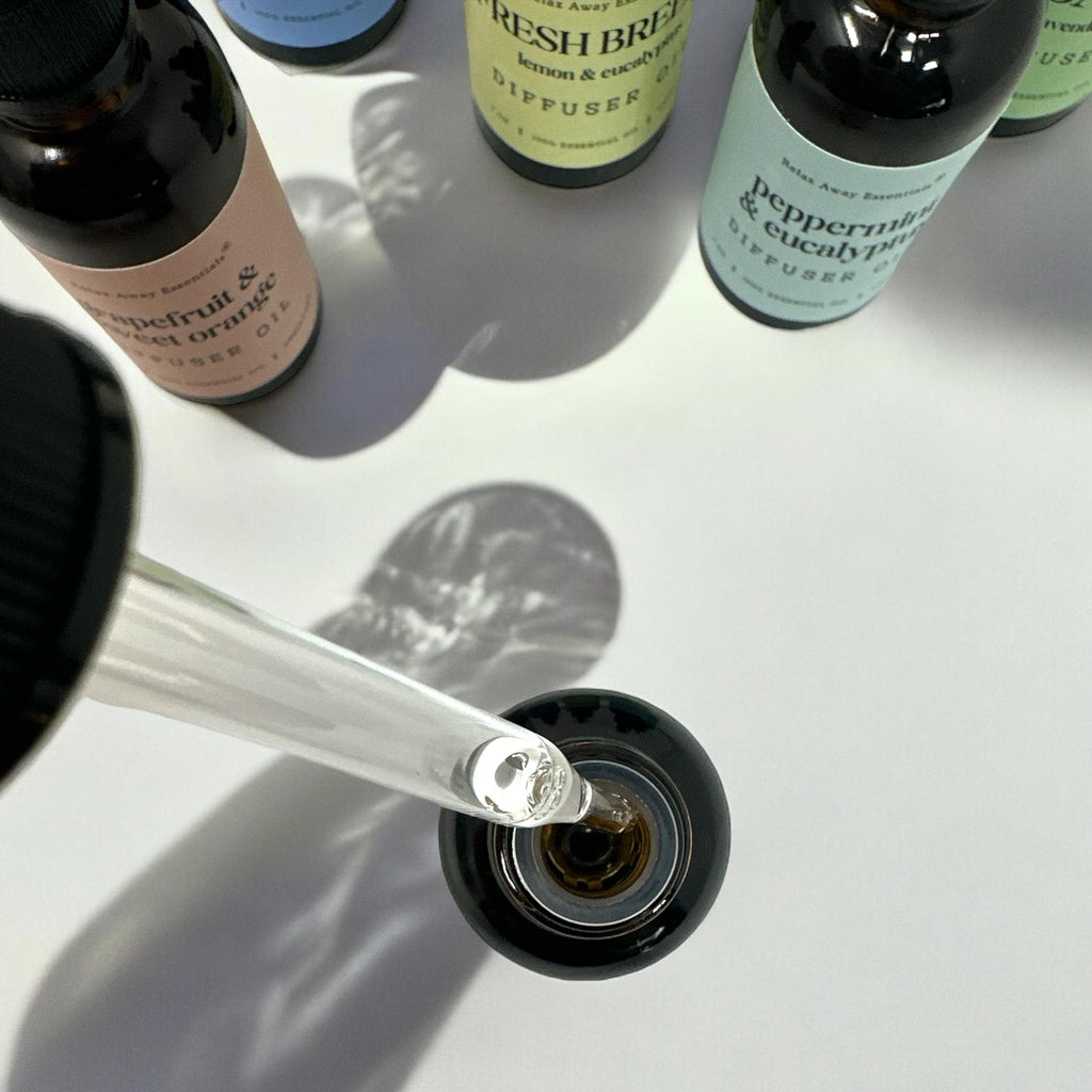 A close-up of a glass dropper filled with clear liquid being dispensed into a small dark bottle. Several diffuser bottles with labels such as peppermint and eucalyptus are visible around it on a white surface with strong shadows.
