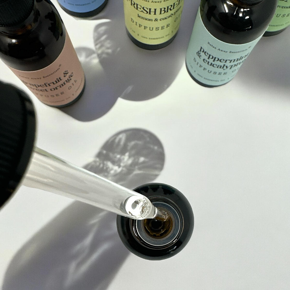 A close-up of a glass dropper filled with clear liquid being dispensed into a small dark bottle. Several diffuser bottles with labels such as peppermint and eucalyptus are visible around it on a white surface with strong shadows.