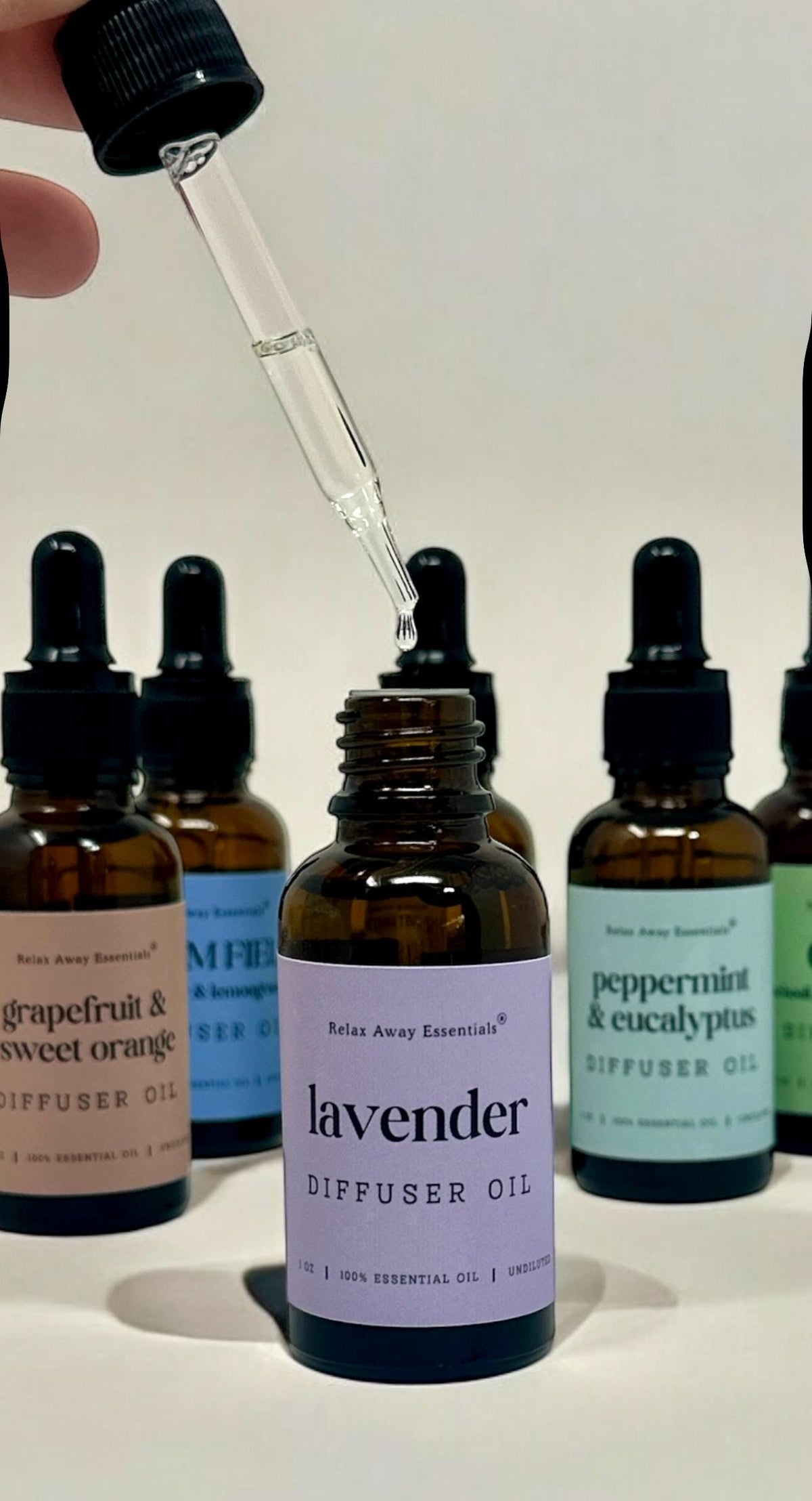 A hand holds a glass dropper above a small amber bottle labeled lavender diffuser oil, with several other essential oil bottles in the background.