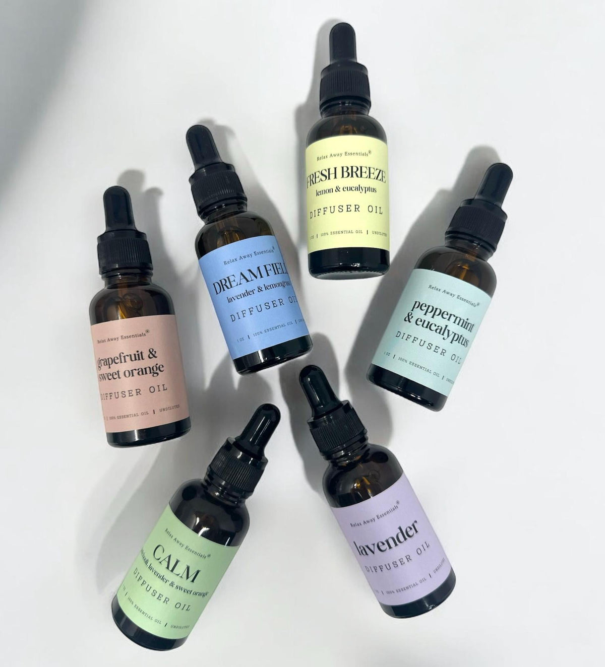 Six amber glass dropper bottles of diffuser oil arranged on a white background, each with a different pastel label: peppermint and citrus, dreamy forest, peppermint and eucalyptus, calm, lavender, and fresh breeze.