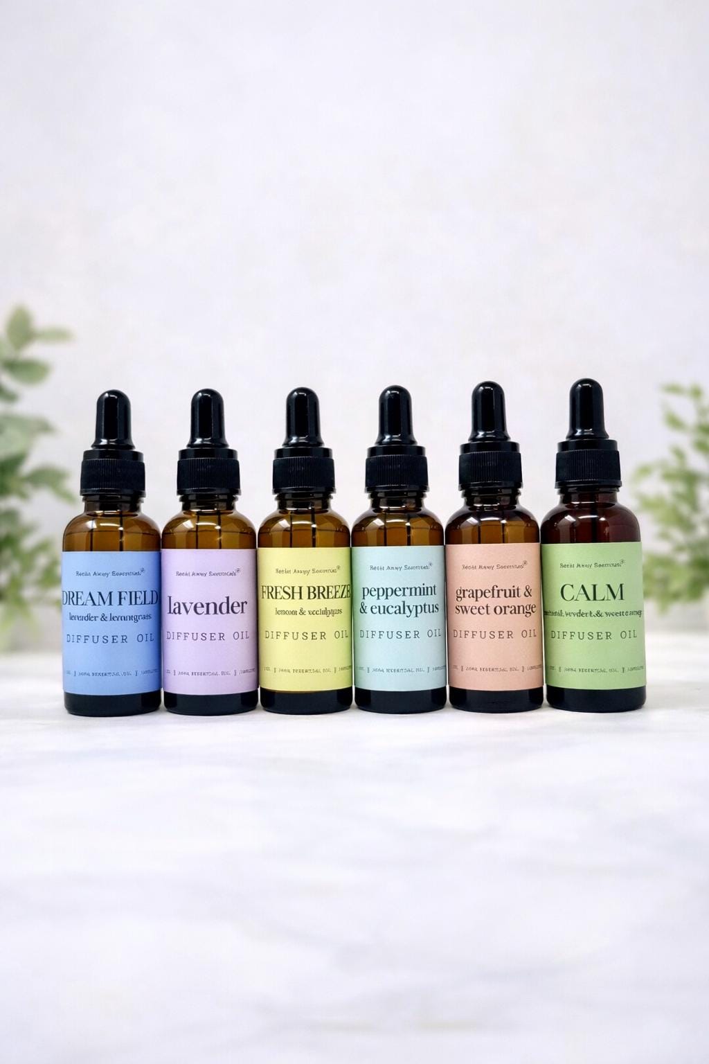 Six amber glass dropper bottles of diffuser oil lined up on a white surface against a soft white background with blurred green leaves. The labels read: Dreamy, Lavender, Fresh Breeze, Peppermint & Eucalyptus, Grapefruit & Sweet Orange, and Calm.