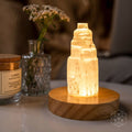 A glowing Selenite step lamp on a round wooden base, set on a tabletop in a softly lit room with blurred decorative bottles and flowers in the background.