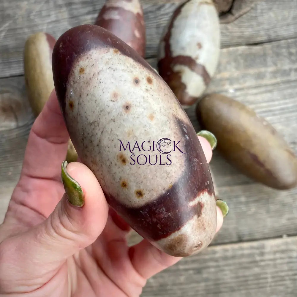 Hand holding a brown and white stone with 'Magick Souls' branding on a wooden surface.