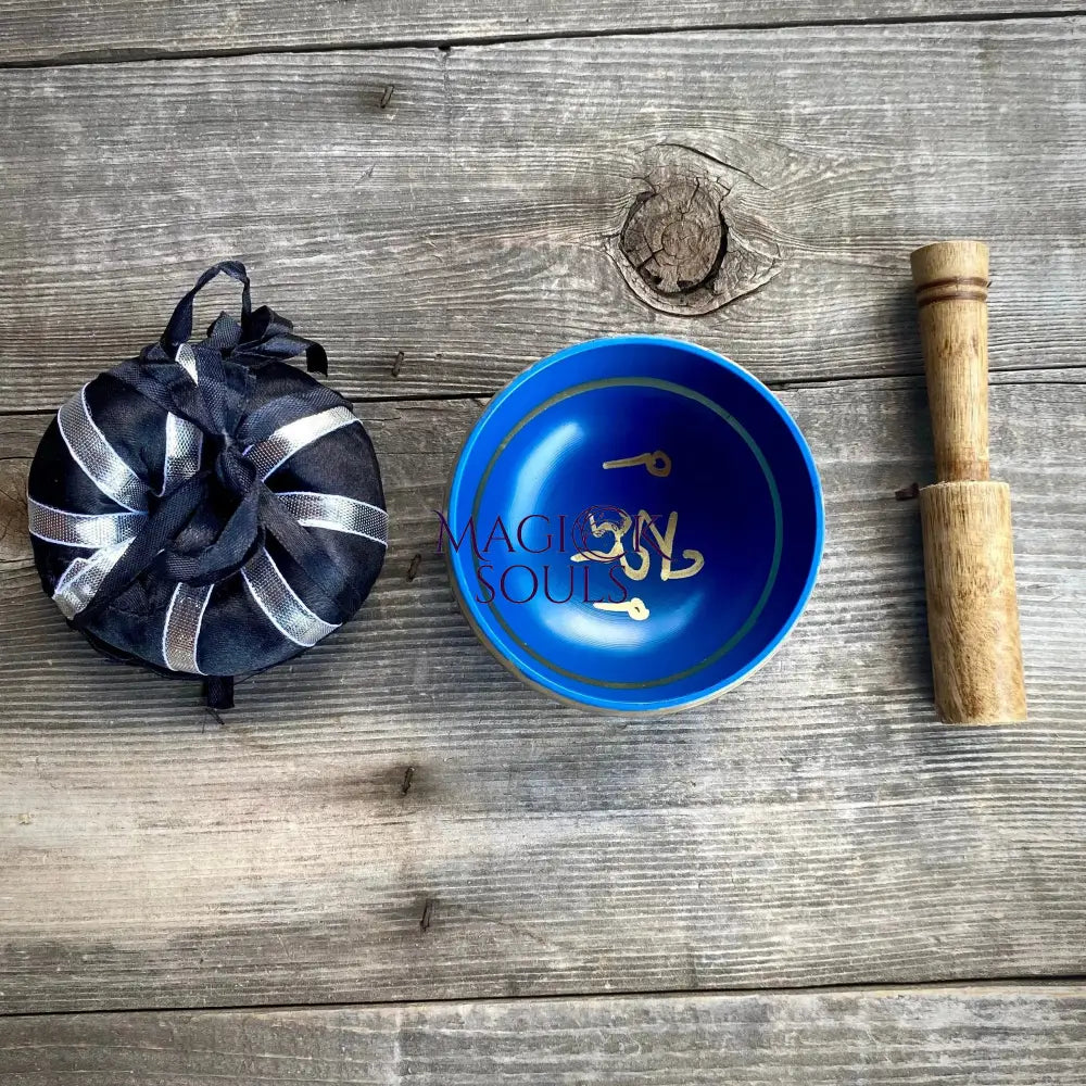 A blue ceramic bowl with gold Arabic calligraphy sits on a rustic gray wooden surface, alongside a black and silver striped fabric pouch and a wooden mallet.