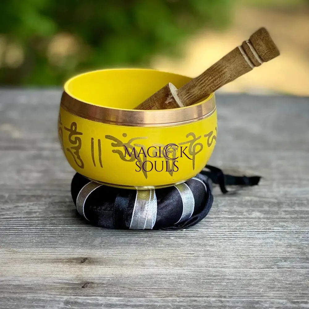 A yellow Tibetan singing bowl with gold-colored script and a wooden mallet resting inside, set on a black and white striped cushion on a weathered wooden surface with a blurred green background. A watermark reading 'MAGICK SOULS' is visible on the bowl.