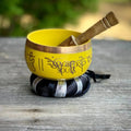 A yellow Tibetan singing bowl with gold-colored script and a wooden mallet resting inside, set on a black and white striped cushion on a weathered wooden surface with a blurred green background. A watermark reading 'MAGICK SOULS' is visible on the bowl.