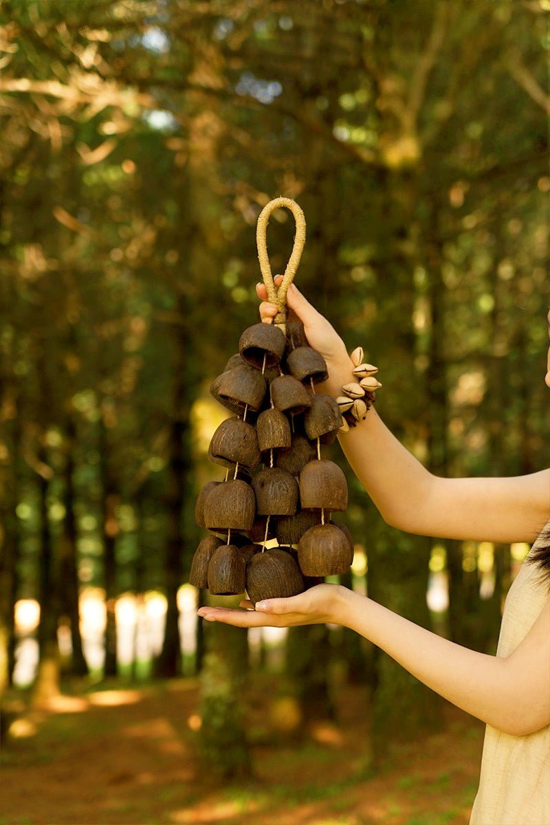 A hand holds a rustic wooden bell wind chime made of stacked coconut shells in a sunlit forest setting.